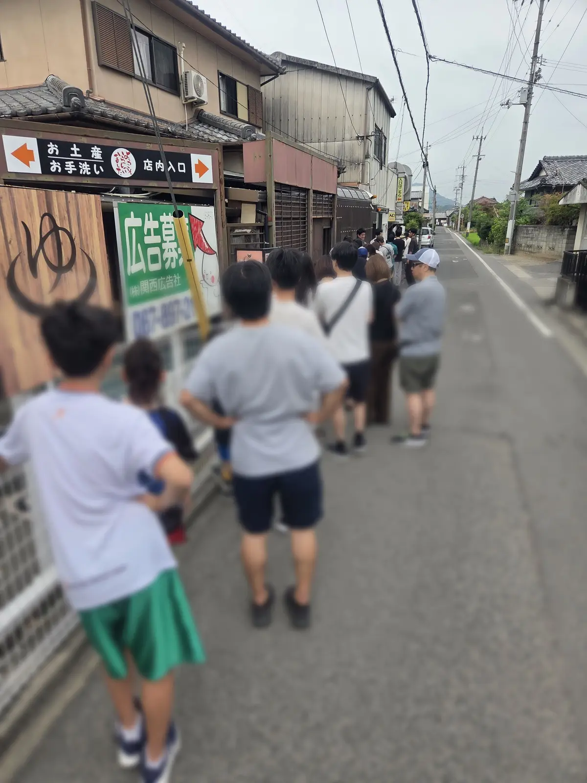 路地裏に面した店先に並ぶ人々の様子。左には「お土産、店内入口」と書かれた看板が見える。灰色の空の下、数人が店を見つめている。