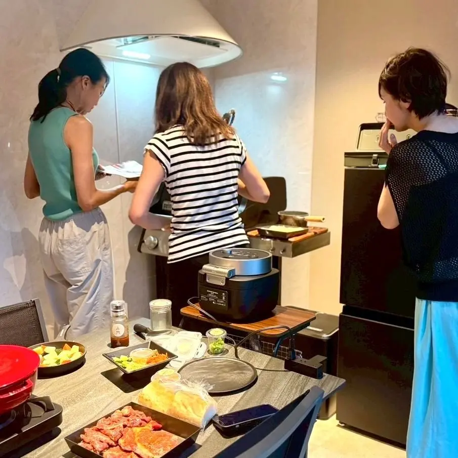 キッチンで3人、ホットプレートを囲んで料理を楽しんでいる様子。ホットプレートの周りには調味料や食材が並び、テーブルには料理が置かれている。