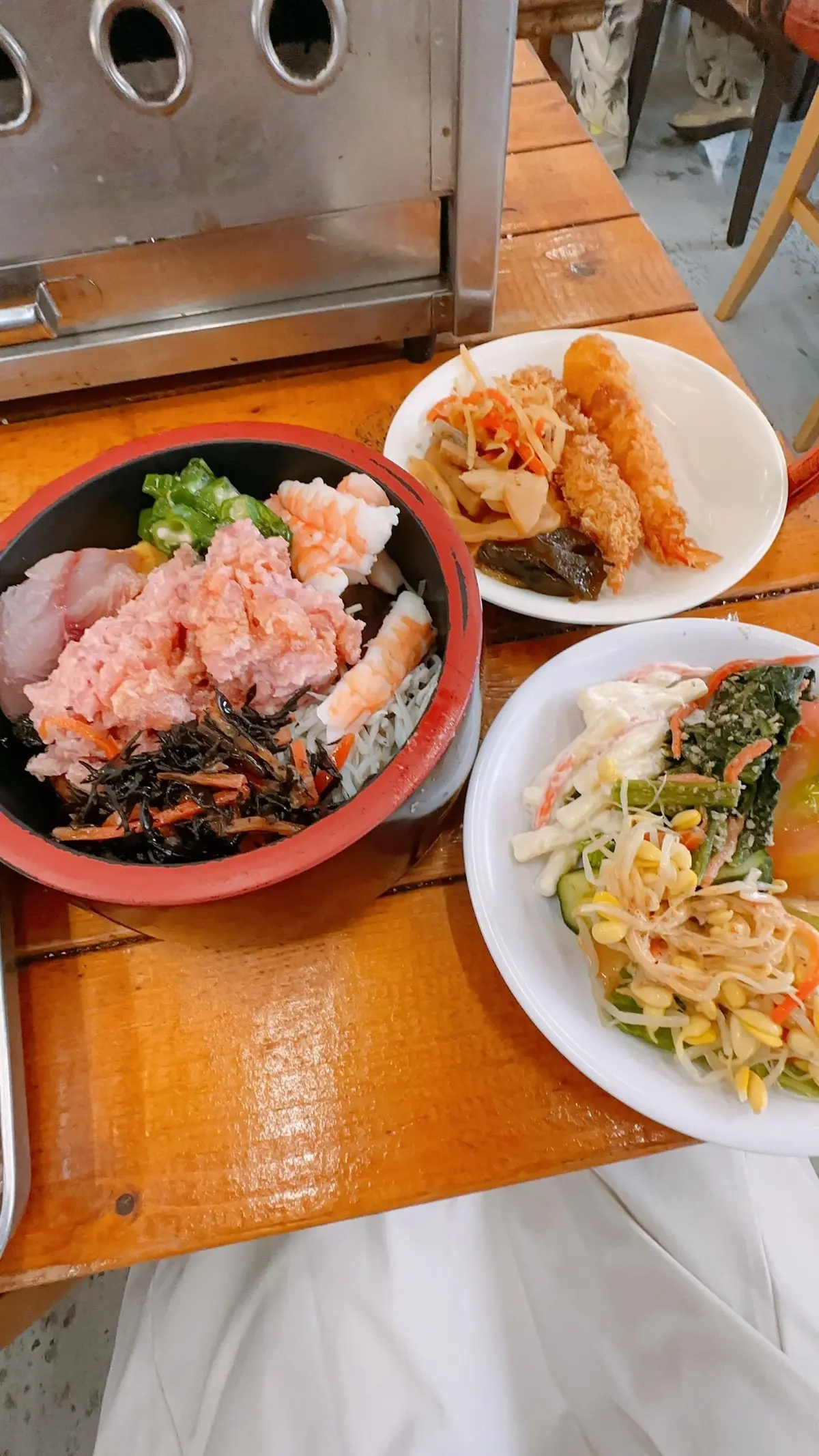 木製のテーブルに置かれた、海鮮丼と小鉢の盛り合わせ。海鮮丼には、刺身、エビ、海苔などが乗っており、小鉢には、野菜とエビのサラダ、魚のフライなどが盛り付けられている。