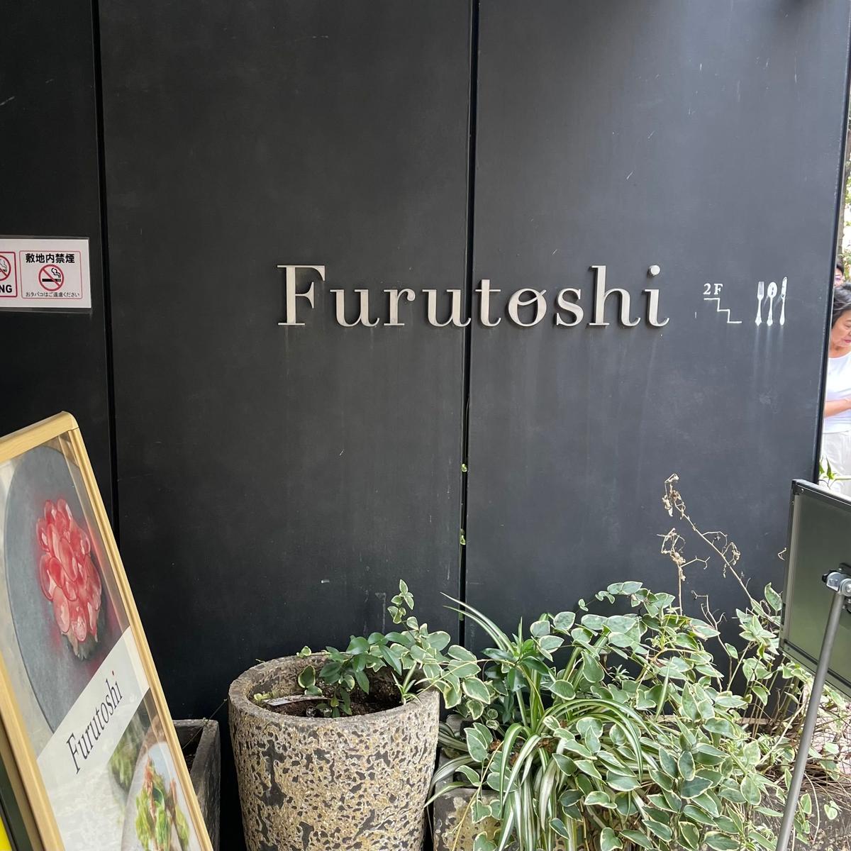 黒いドアに白で「Furutoshi」と表記された看板。2階を示す「2F」の文字と、植物のイラストが添えられている。ドアの横には、植物を入れた鉢と、Furutoshiのロゴが入った立て看板が置かれている。