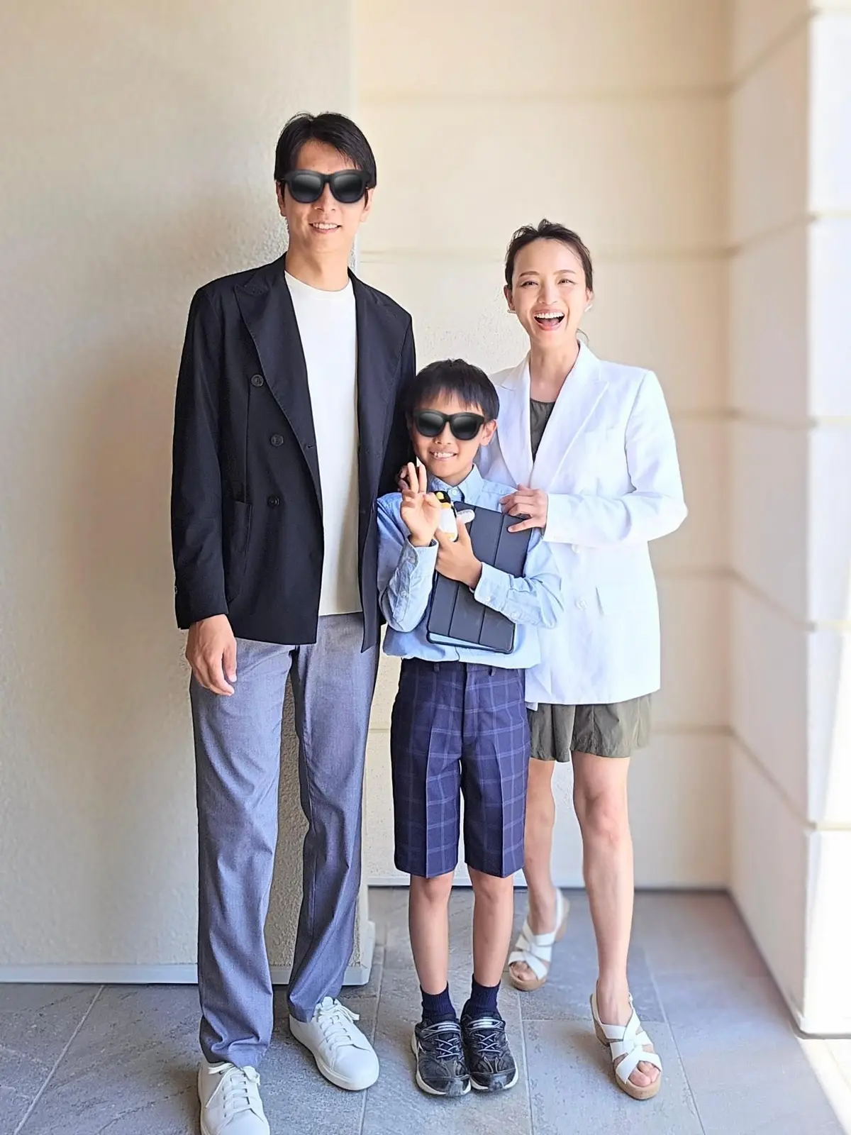 笑顔の家族3人が屋外で記念撮影をしている。左は黒いジャケットに白いシャツ、右はライトブルーのジャケットにシャツ姿の女性。中央は制服姿の少年で、黒縁のサングラスをかけている。背景にはベージュ色の壁がある。