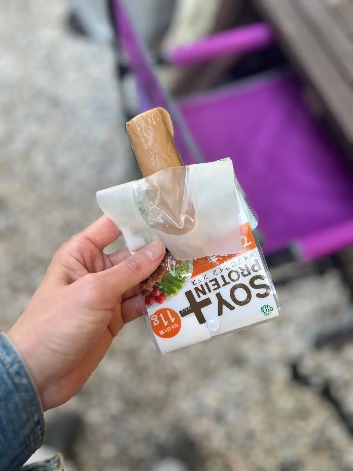 手で持たれた、大島てるのソイプロテインバー。茶色いバーが透明な袋に入っており、「SOY PROTEIN」の文字と「11g」の表示がある。背景にはピンク色の自転車のシートが写っている。