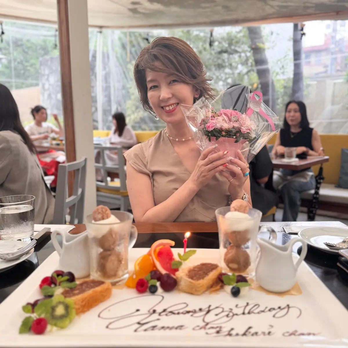 レストランで誕生日のお祝いを受ける女性。笑顔でピンクの花束を持ち、テーブルのパンケーキとフルーツを背景に写る。周囲には他の客とスタッフが見える。