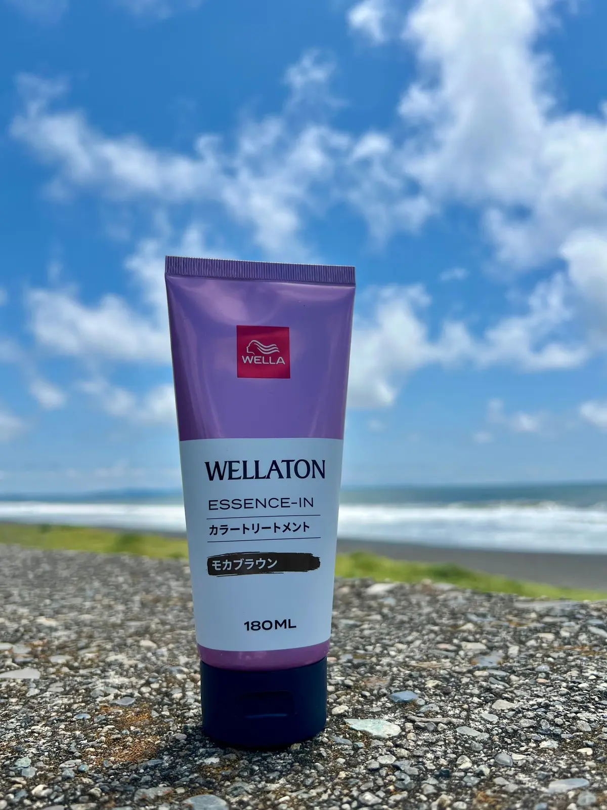 海岸を背景に置かれた、Wella（ウェラ）のヘアケア製品「Wellaton エッセンスイン カラートリートメントメント」の180mlチューブ。白地に紫色のキャップ、茶色の底部分。