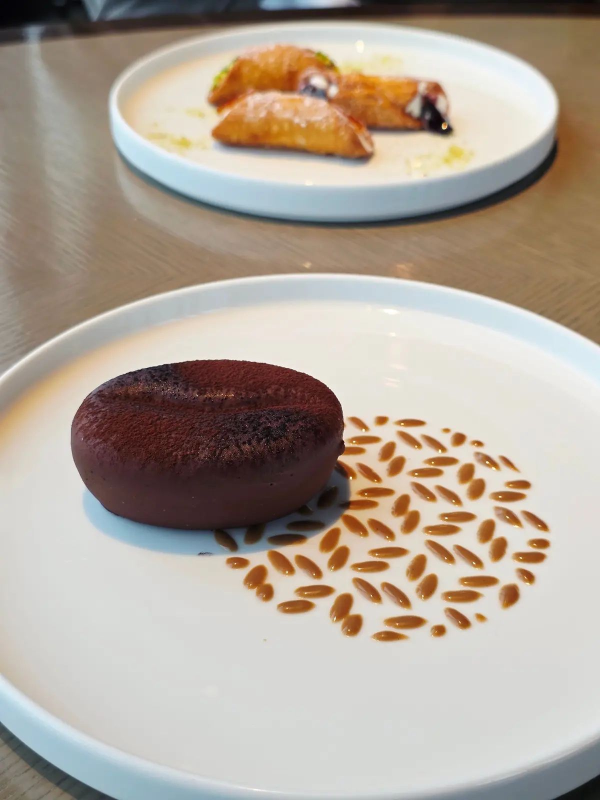 白磁の皿に、丸いチョコレートケーキと、キャラメルソースで描かれた花模様。背景には、揚げ菓子が乗った皿がぼやけて写っている。