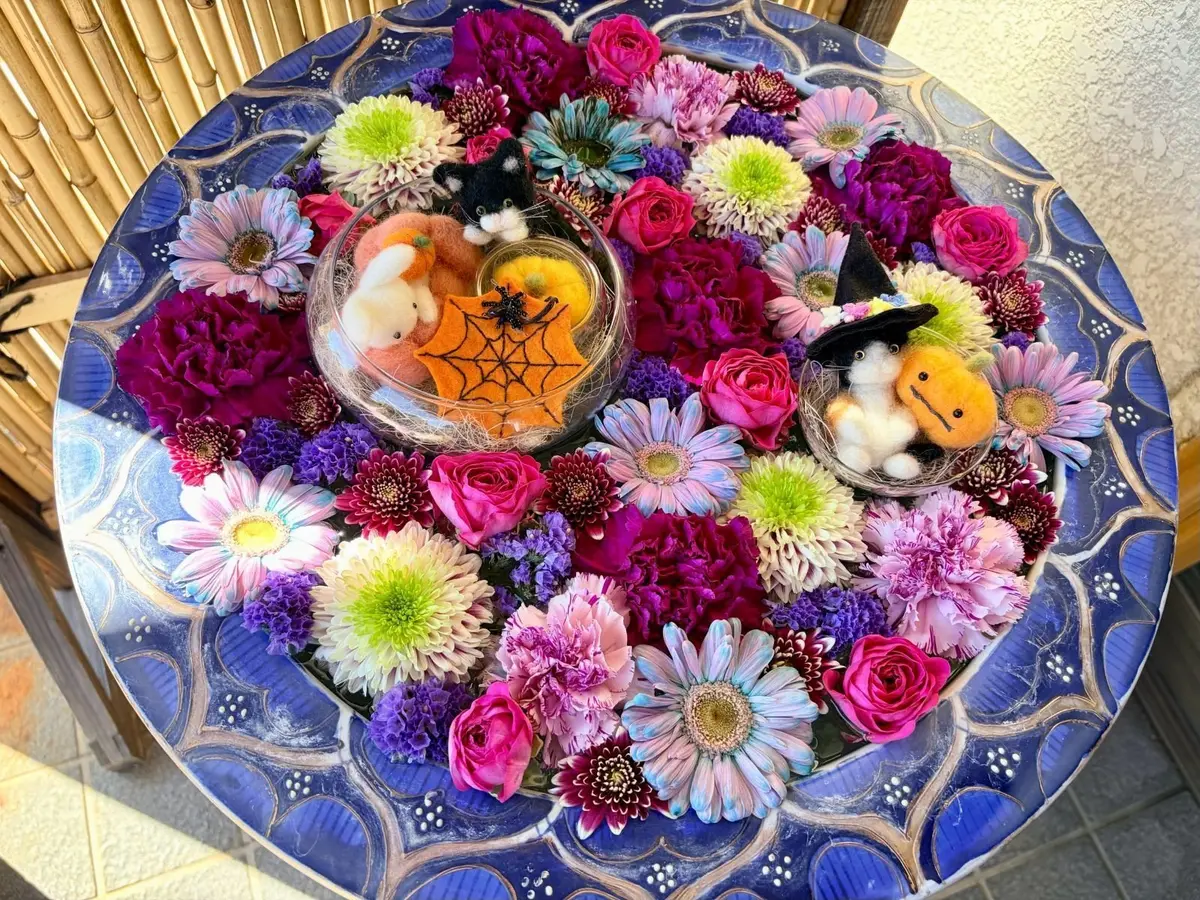 丸いテーブルに、紫、ピンク、白、緑の様々な花々がふんだんに飾られたハロウィンアレンジメント。中央には、蜘蛛の巣と小さな置物が飾られたガラスの器が2つ置かれている。