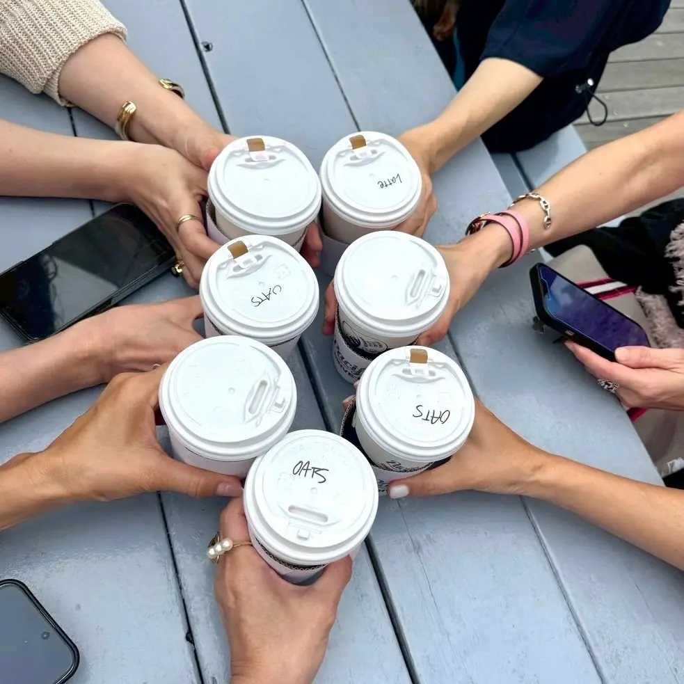 パシフィックドライブインのコーヒーカップ。7人で乾杯！複数の人がテーブルに並んだコーヒーカップを囲み、ラベルが書かれたカップをそれぞれ持ち、スマートフォンも置かれている