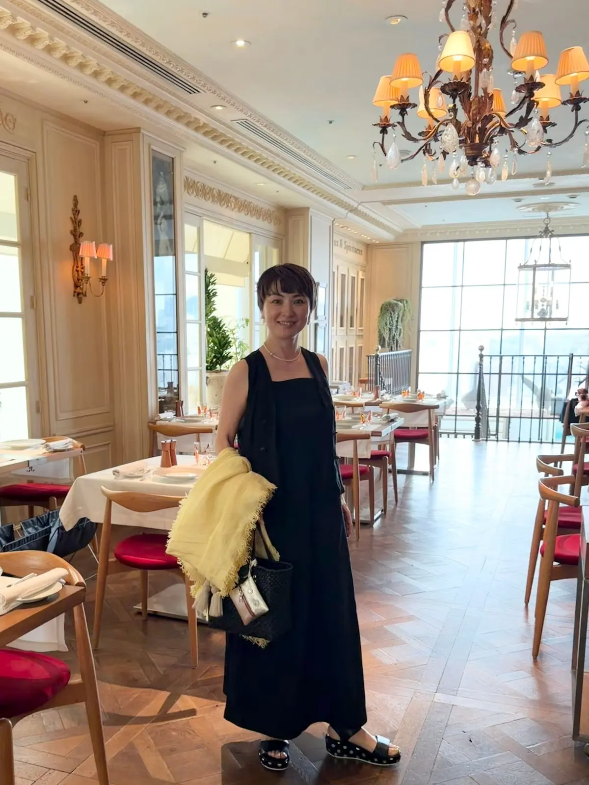 黒いロングドレスを着た女性が、明るいレストランの中で立っている。背景にはシャンデリアと窓からの景色が見える。女性は黄色いショールと黒いバッグを持っている。
