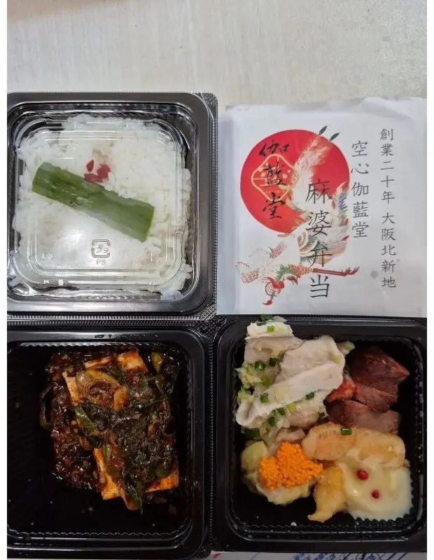 白米と緑色の野菜、黒いソースで炒めた野菜、白身魚と野菜、赤身の肉が盛り付けられた中華弁当。赤いロゴ入りの紙が添えられている。