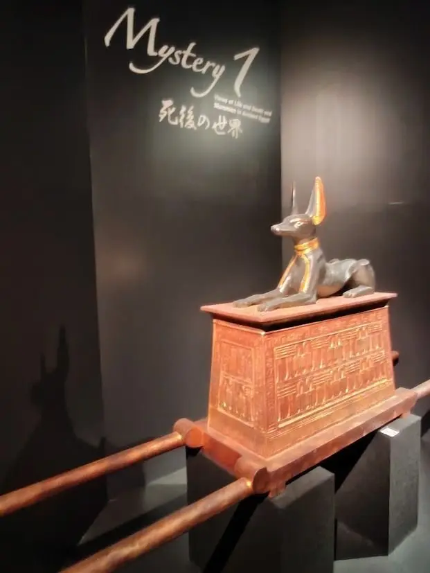 展示ケースの上の、黒い犬の頭をした像。像は、金色の装飾が施された、赤褐色の箱の上に座っている。背景には「Mystery 1」の文字が書かれた黒い壁。