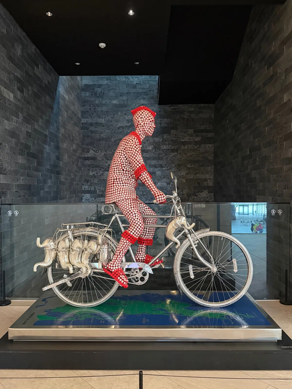 赤いドット模様の服を着た人物が、銀色の自転車に乗り、展示ケースの中に置かれている。