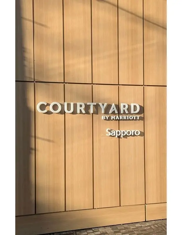 木目調の壁面に、銀色の「COURTYARD BY MARRIOTT Sapporo」のロゴがはめ込まれている外観写真。右側に影が映っている。