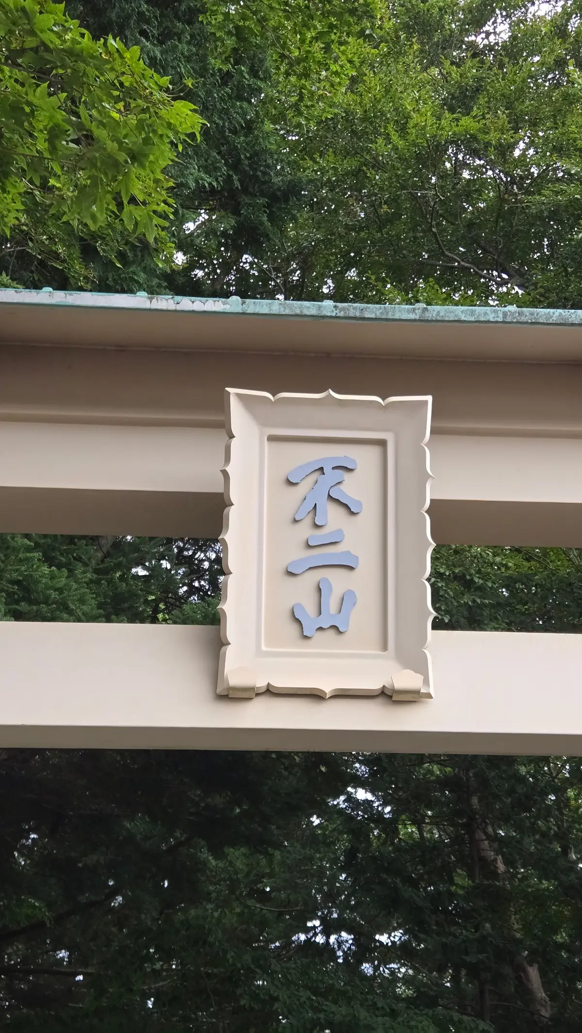 白地に青色の額縁に収められた、縦書きの漢字「不二山」の標識。背景には緑豊かな木々が見える。