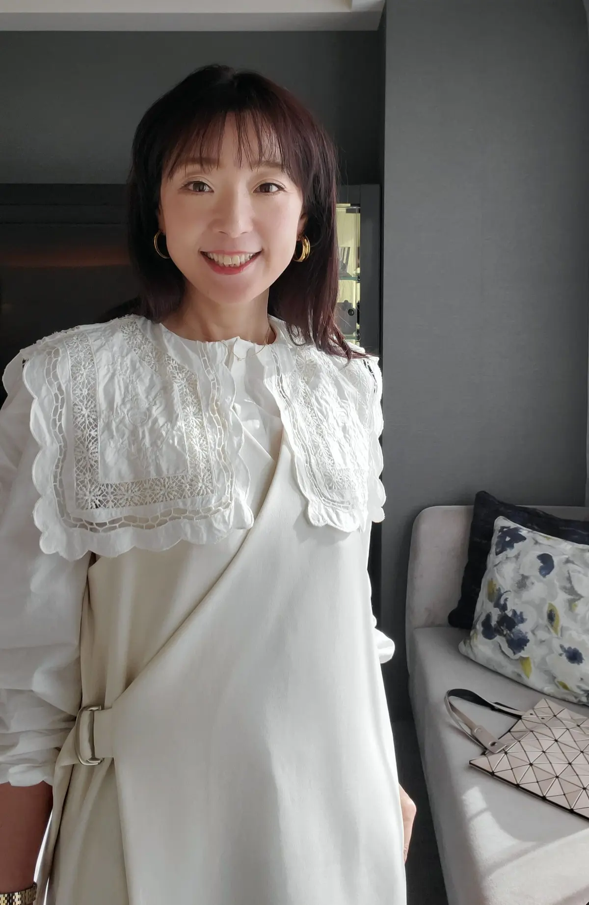 女性が白いレースのブラウスを着て、笑顔でカメラに向かって立っている。背景にはグレーの大型ソファがあり、青い花模様のクッションと幾何学模様のトートバッグが置かれている。
