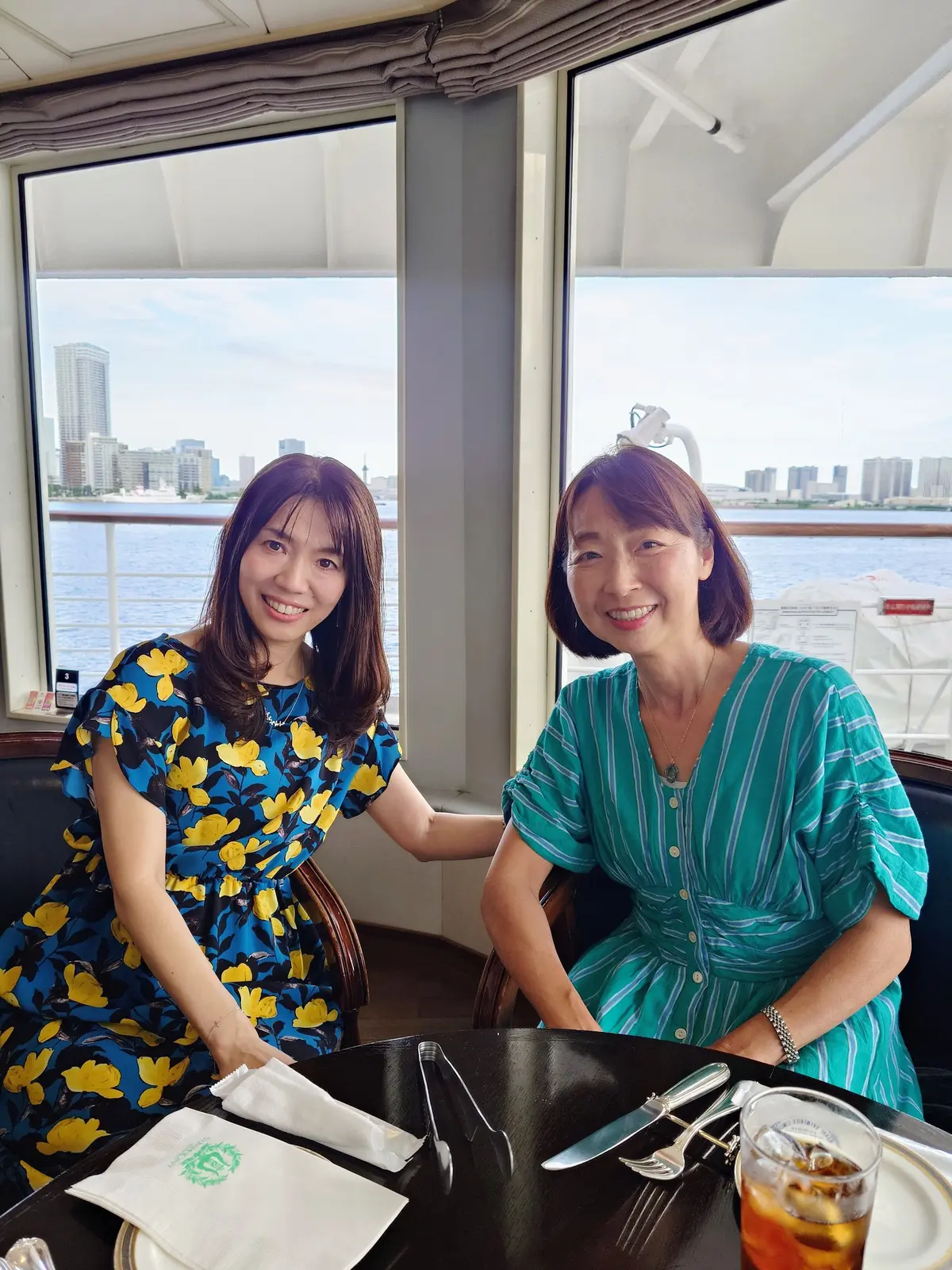 窓から水辺の景色を望む、笑顔の親子。左は黄色の花柄ワンピースを着た若い女性、右は水色のストライプのワンピースを着た女性。テーブルには飲み物とカトラリーが置かれている。