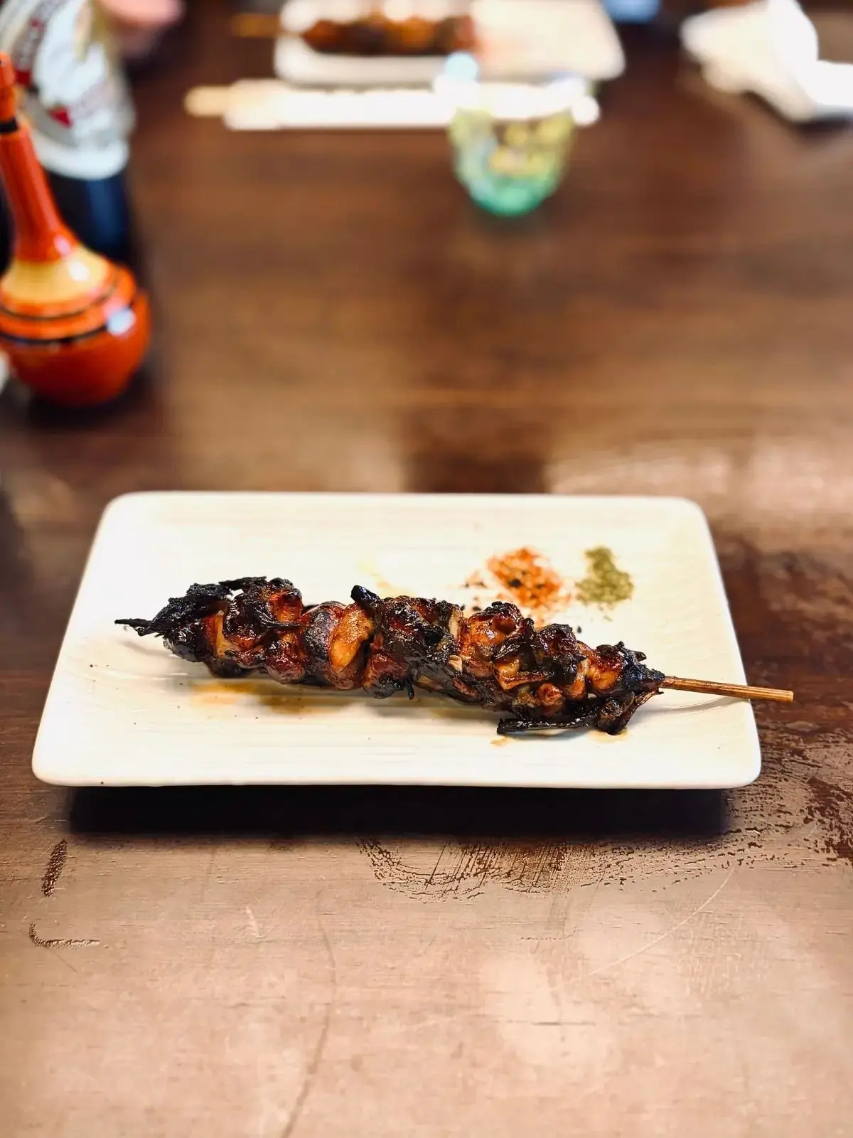 串に刺さった照り焼きチキン。表面は焦げ目がつき、赤唐辛子と緑色の粉末が添えられている。白い長方形の皿に盛り付けられ、木製のテーブルに置かれている。