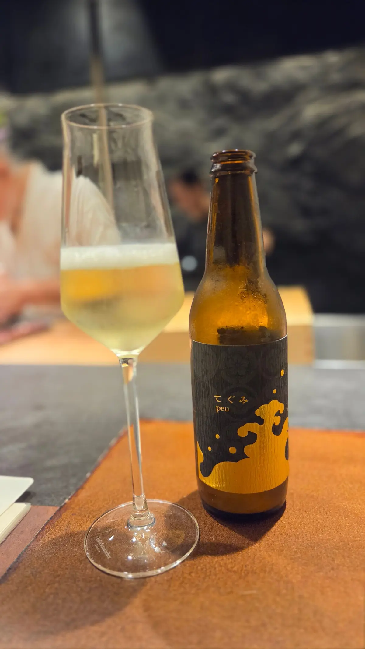 テーブルに置かれた、ツクツクpeuのビール瓶とグラス。グラスには淡い色のビールが注がれており、瓶は茶色のラベルに金色の模様が描かれている。
