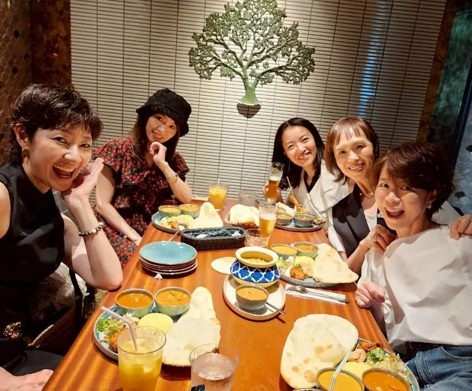 木のテーブルに多様な料理と飲み物が並び、5人の女性が笑顔で楽しんでいる。背景には窓と人工の木の装飾がある。