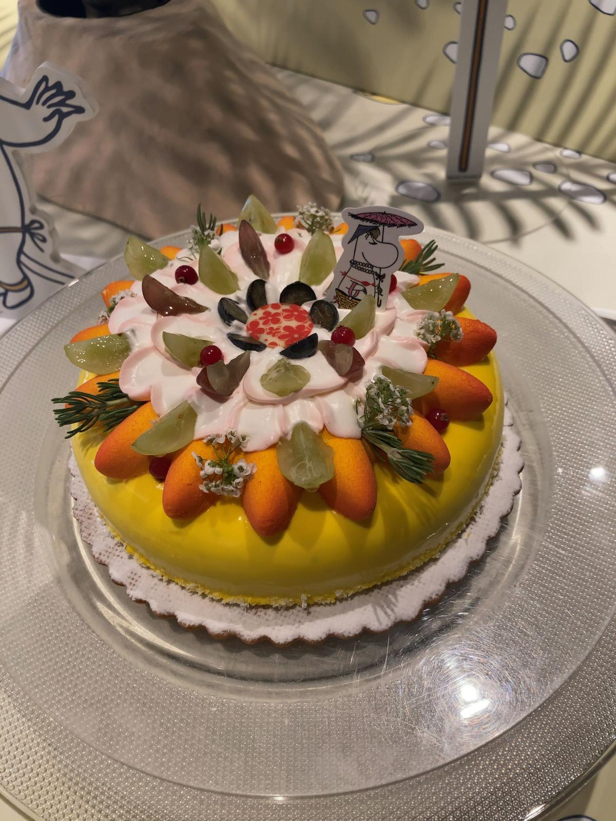 黄色いケーキにクリーム、フルーツ、グリーンの装飾がトッピングされ、小さな紙の旗が立っている