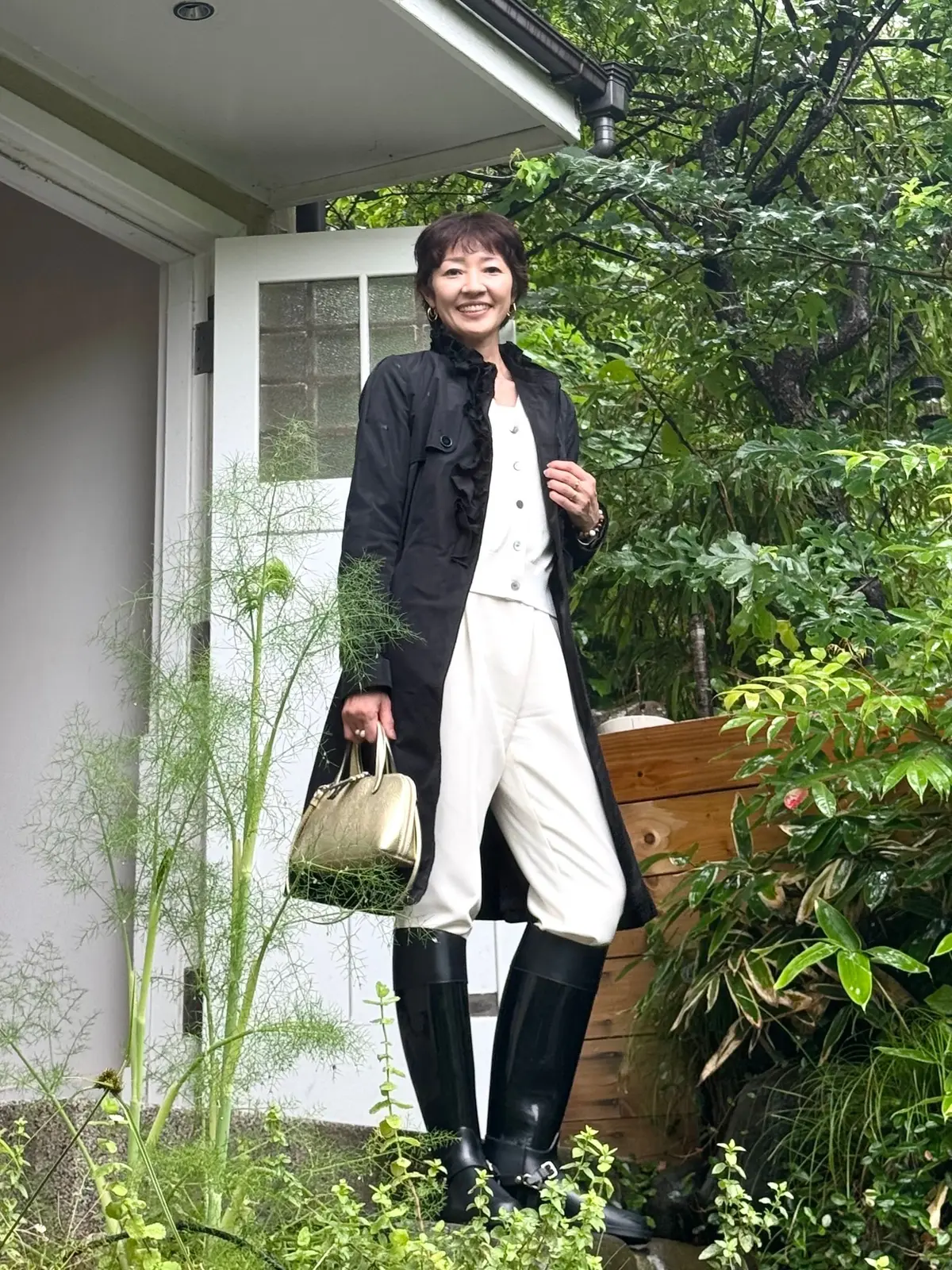 女性が白い家屋の前に立っており、黒の長コートとベージュのワイドパンツを着用しています。黒のブーツを履き、右手には金色のハンドバッグを持っています。周囲には緑豊かな植物が広がっています。