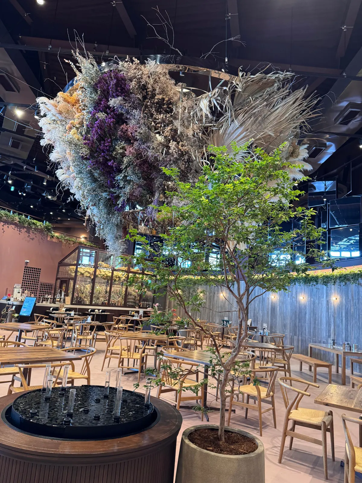 木製のテーブルと椅子が並ぶレストランの内装で、中央に小さな木が植えられ、大きな花の装飾が天井から吊るされている
