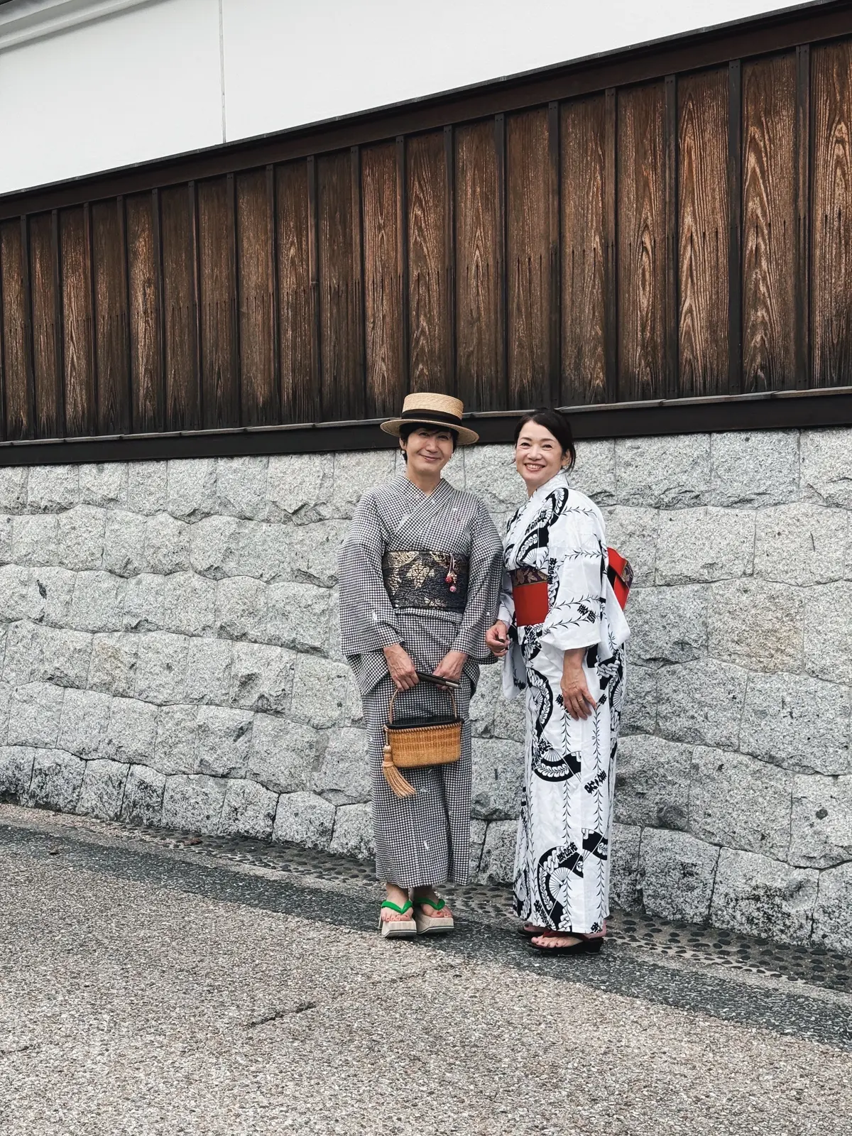 石造りの壁の前に立つ2人の女性。左の女性はギンガムチェックの着物と帽子、右の女性は白黒の柄の着物を着用。それぞれ腰に小物を付けている。