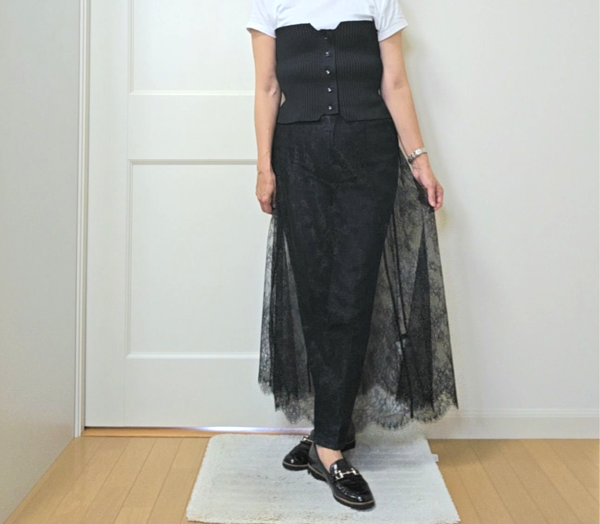 白いTシャツの上に黒いコルセット、黒いレースのスカートとタイツを着用した女性が立っている全身写真。黒いローファーを履き、左手首にブレスレットをしている。背景は白いドアと木製の床。