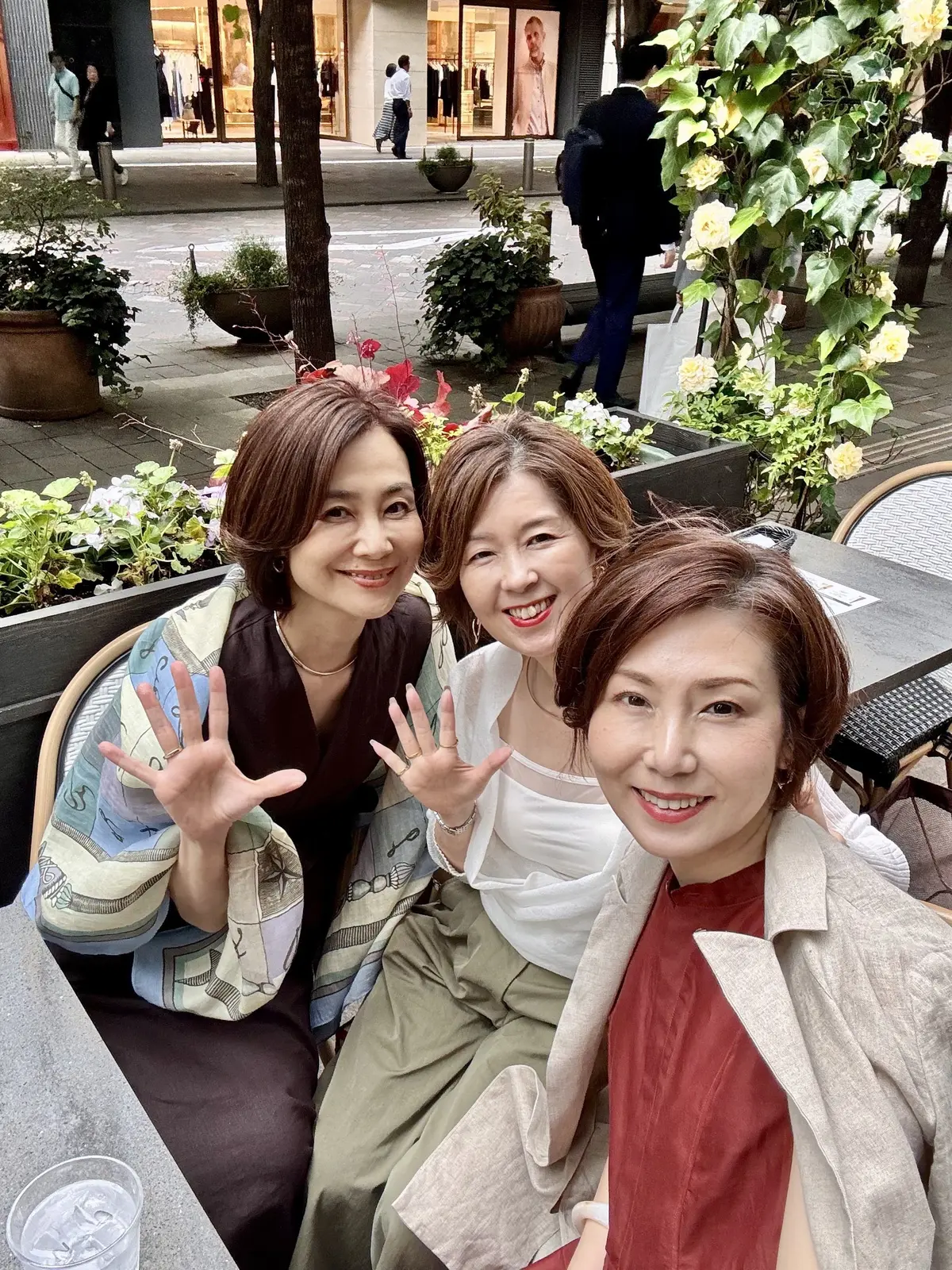 3人の女性が屋外のカフェで笑顔を見せ、手を振っているセルフィー写真。左から茶髪の女性、中央は白いシャツを着た女性、右は赤色のトップスを着た女性。背景には植栽や歩道、店舗が見える。