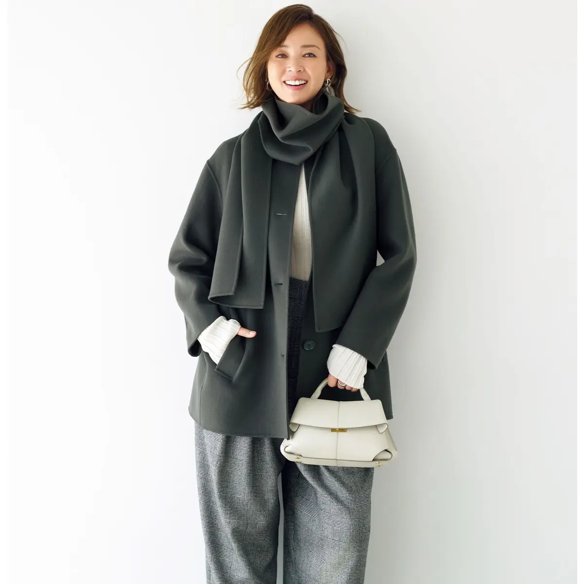 緑色の無地コートに、グレーのチェック柄パンツを合わせた女性。オフホワイトのタートルネックセーターを着用し、緑色のマフラーを巻いている。手には白いハンドバッグを持つ。笑顔でこちらを見ている。