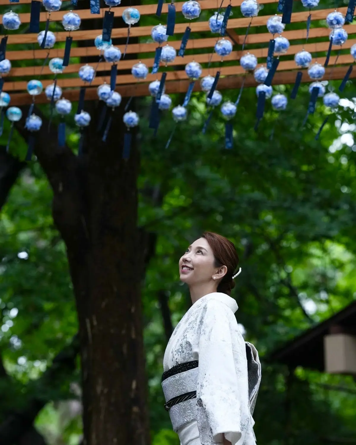 木製の屋根に青い花飾りが吊るされ、白い着物を着た女性が笑顔で振り返っている