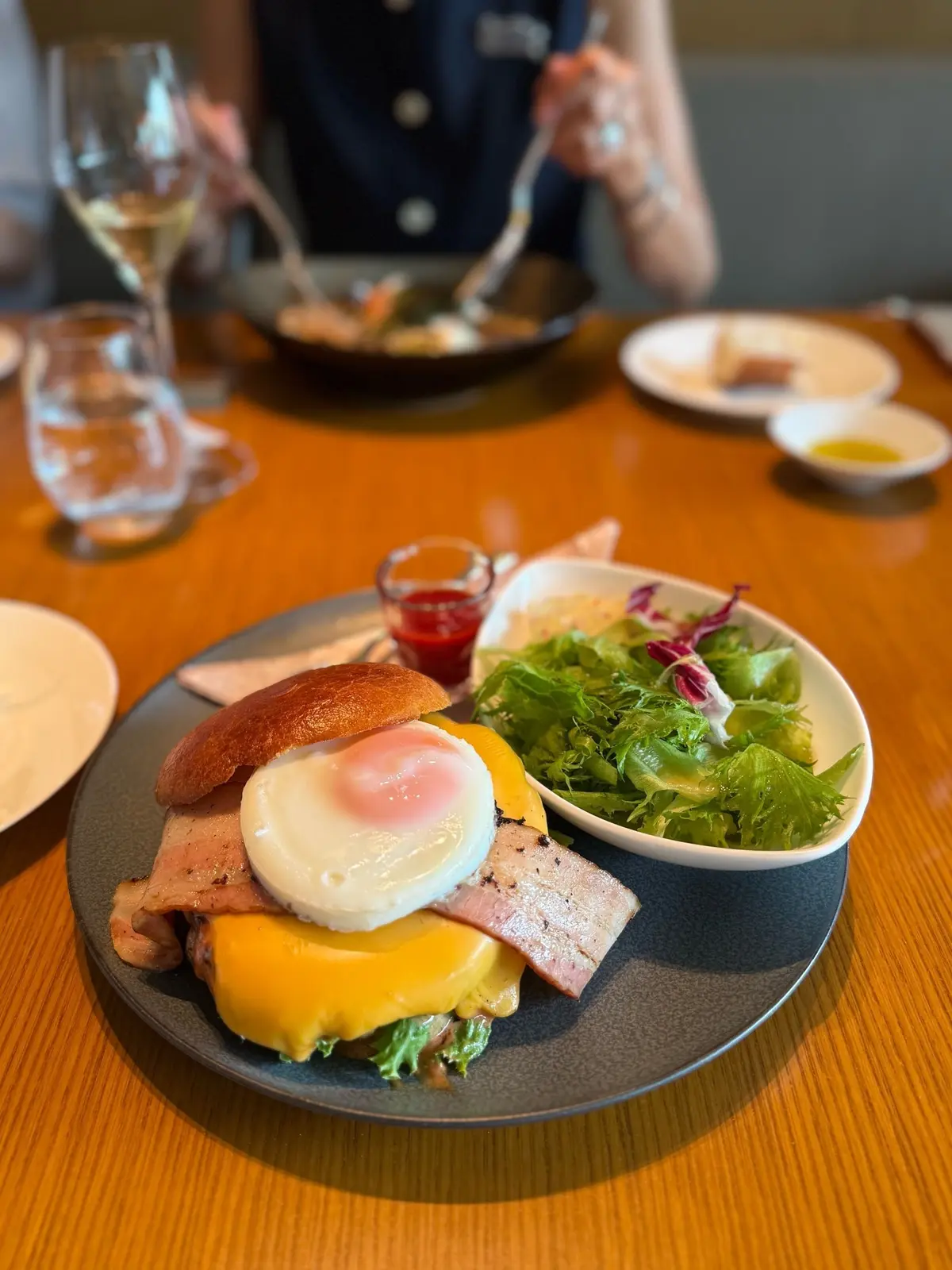 木製のテーブルに置かれた、グレーの皿の上のハンバーガー。バンズにベーコン、チーズ、目玉焼きが挟まれており、横には小さなボウルに入ったグリーンサラダと赤いソースが添えられている。背景にはワイングラスと、他の料理が写っている。