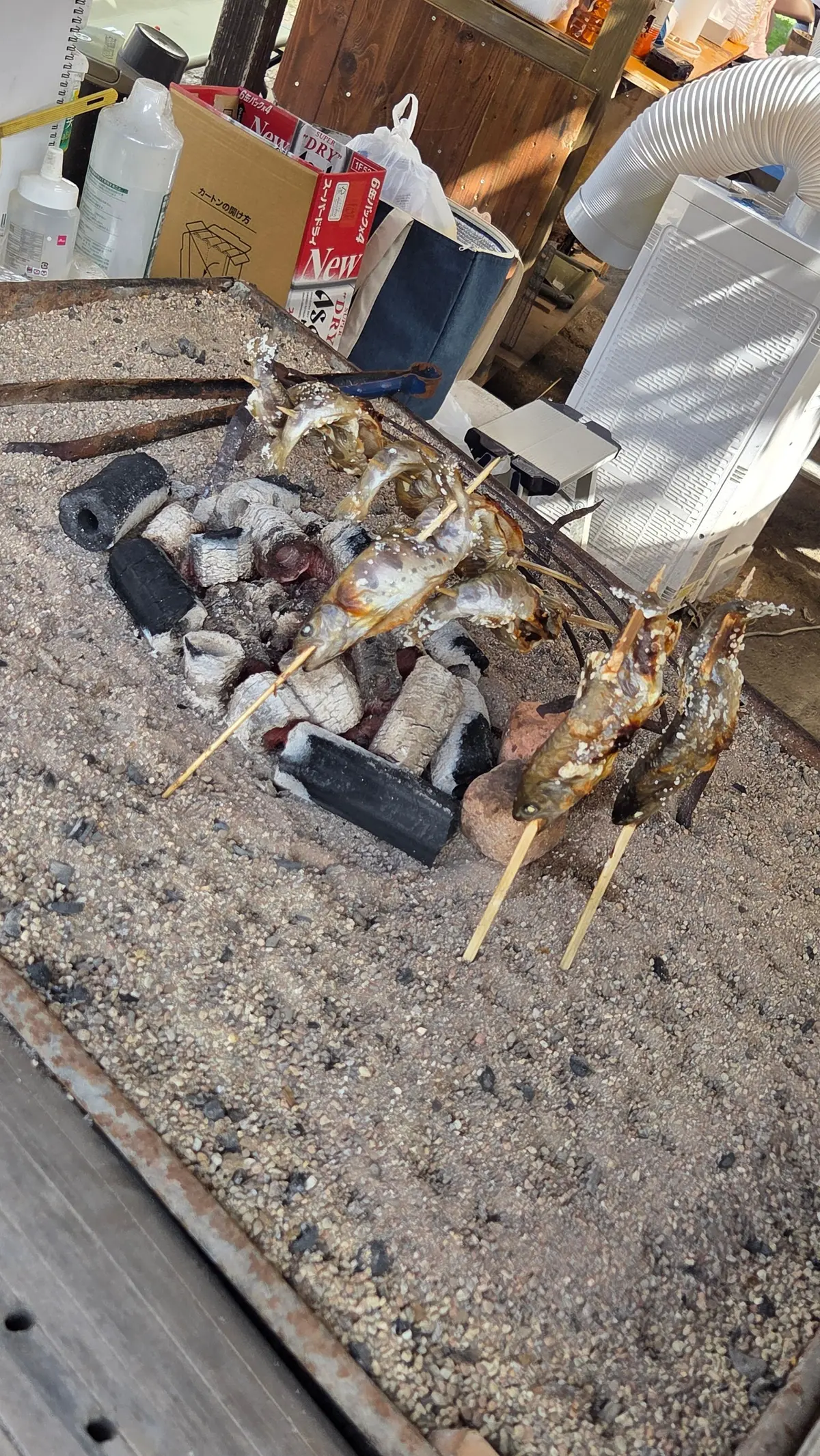 砂利敷きの屋外で、魚を串焼きにしている様子。黒い炭の上に魚が並び、棒で支えられている。背景には、箱や椅子、扇風機などが置かれている。