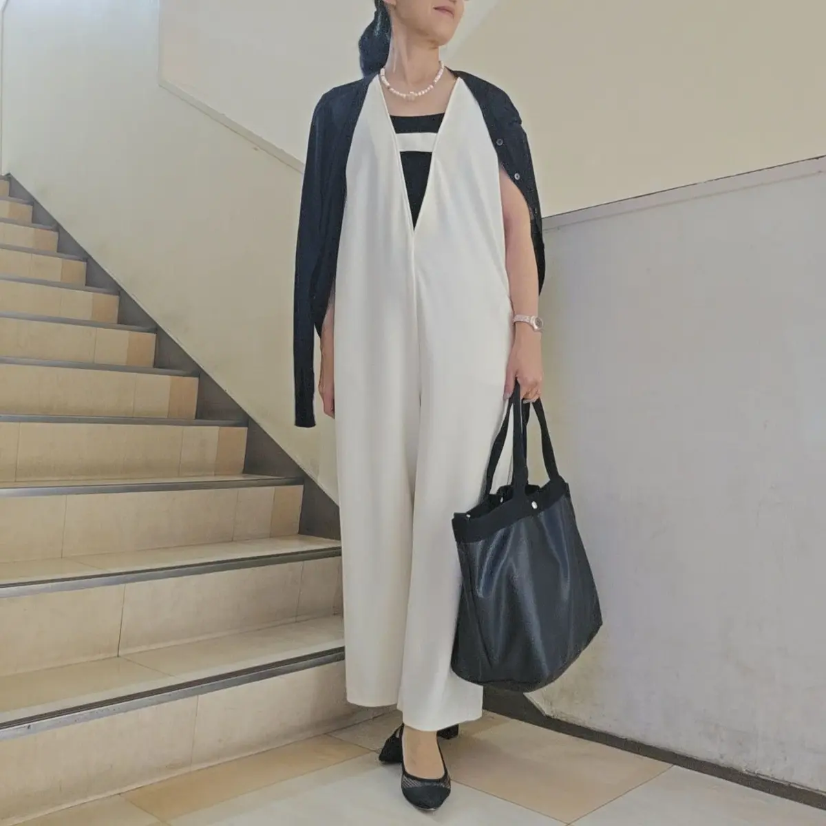 黒いカーディガン、白黒ボーダーのトップス、白いオーバーオールを着用した女性。黒いハンドバッグを持ち、黒いパンプスを履いている。背景には階段がある。