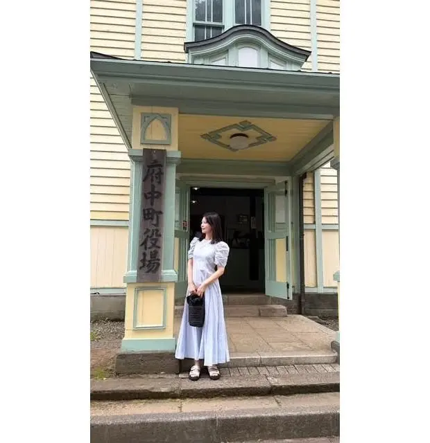 伝統的な和風建築の玄関前に立つ女性。薄青のロングドレスを着て、黒い手提げ袋を持ち、玄関の石段に足を踏み出し、建物の看板を眺めている。