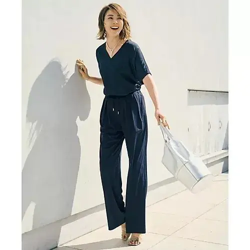 濃紺のワイドパンツとトップスがセットになった服を着た女性。Vネックで、腰にベルトあり。白いバッグを持ち、笑顔でこちらを見ている。背景は白い壁。