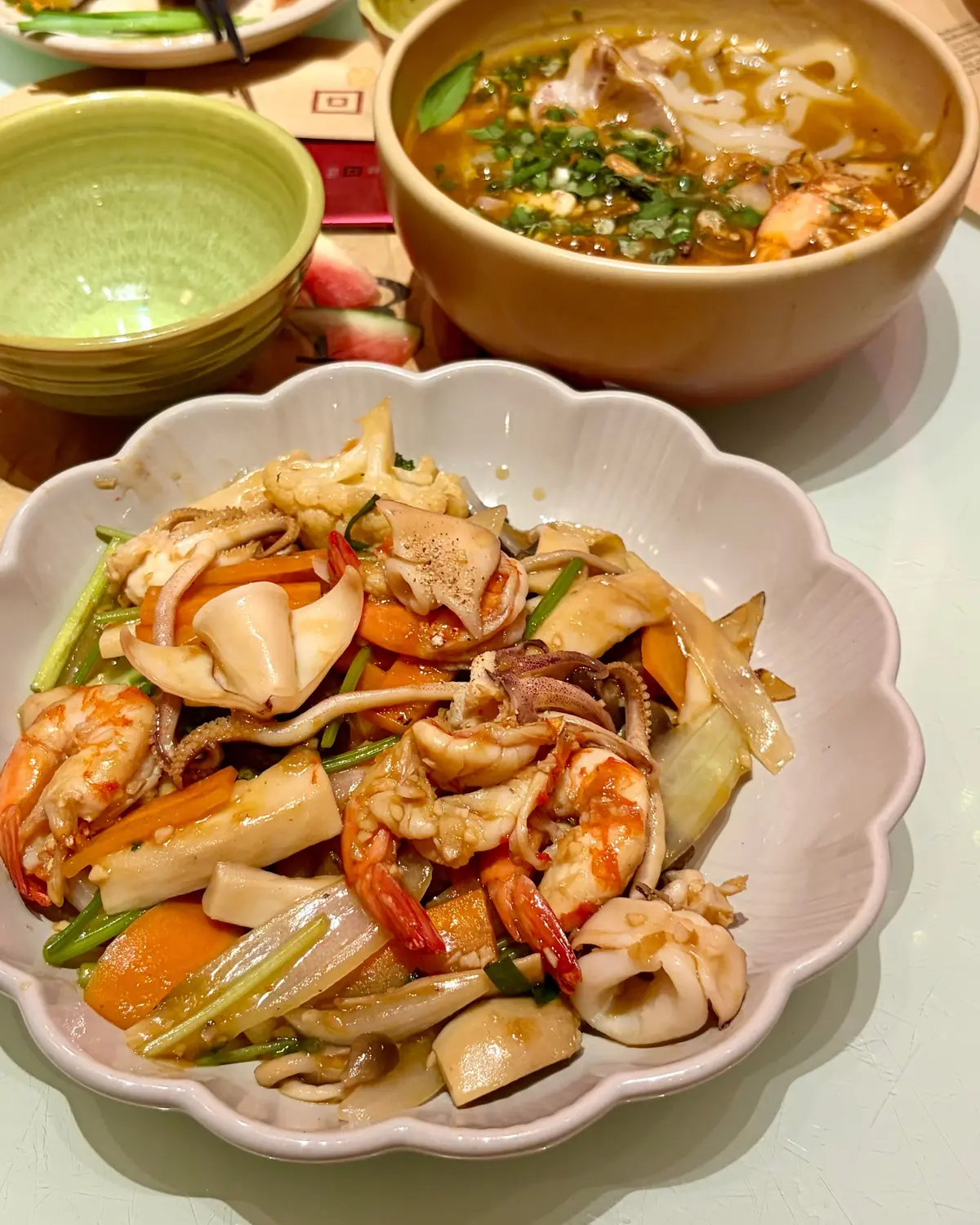 エビやキノコ、野菜が盛りされた白い波状の皿と、緑の汁物が入った黄色いボウルが並ぶ