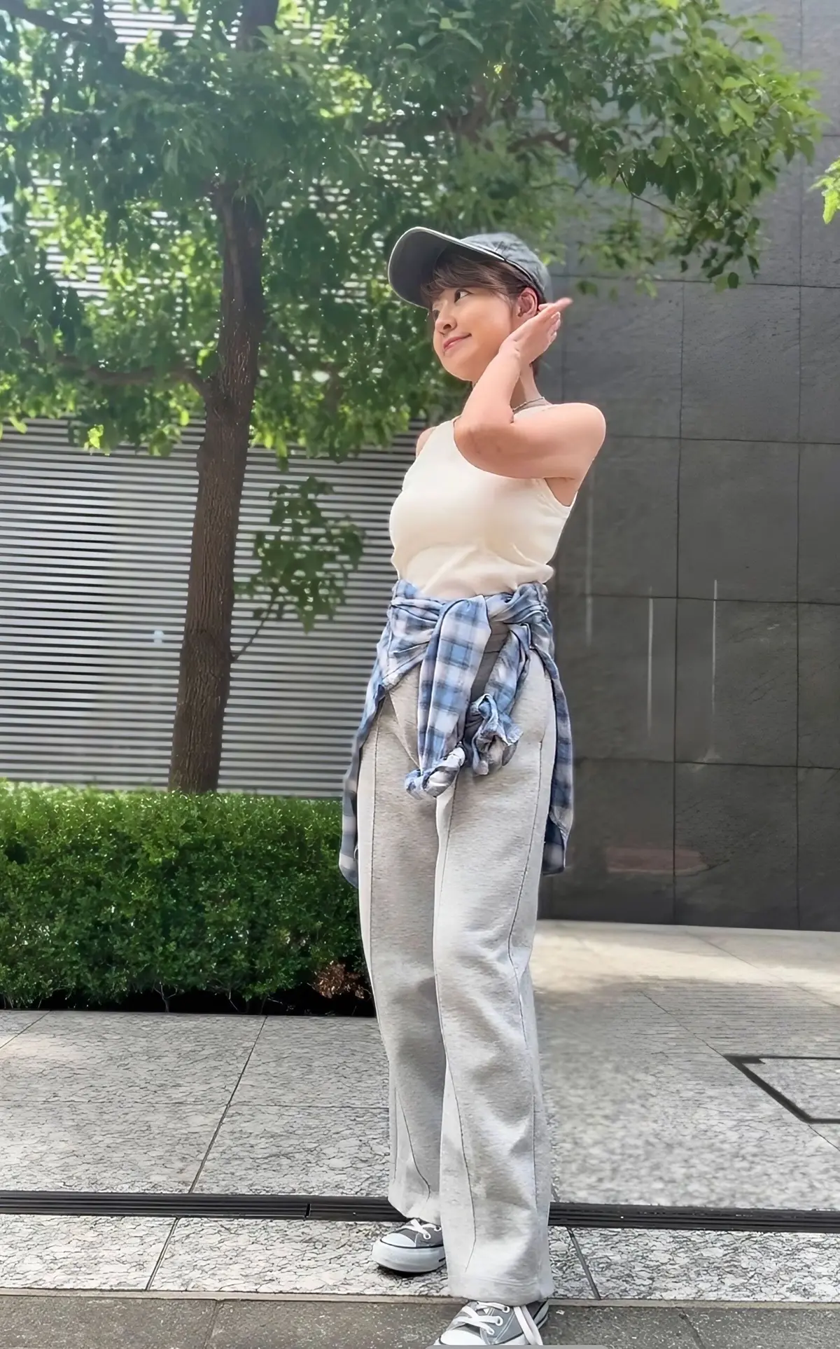 女性がグレーのキャップをかぶり、ベージュのタンクトップとグレーのワイドパンツを着て、左手を額に当ててポーズしています。背景には緑の木や低木があり、コンクリート敷きの屋外空間です。