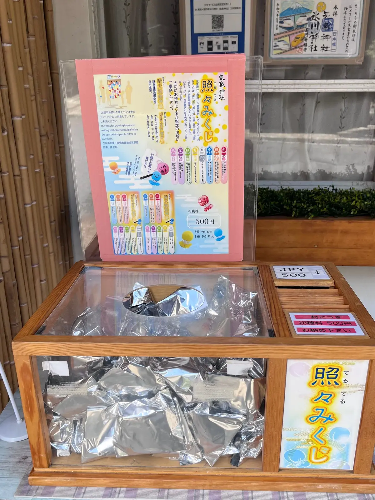 ピンクと黄色のポップなデザインの案内板と、金色の包みに入ったお菓子が並んだ木製の陳列ケース。右には料金表示の立て札と、お菓子の名前が書かれた黄色の看板がある。背景には石造りの壁と竹の柵が見える。