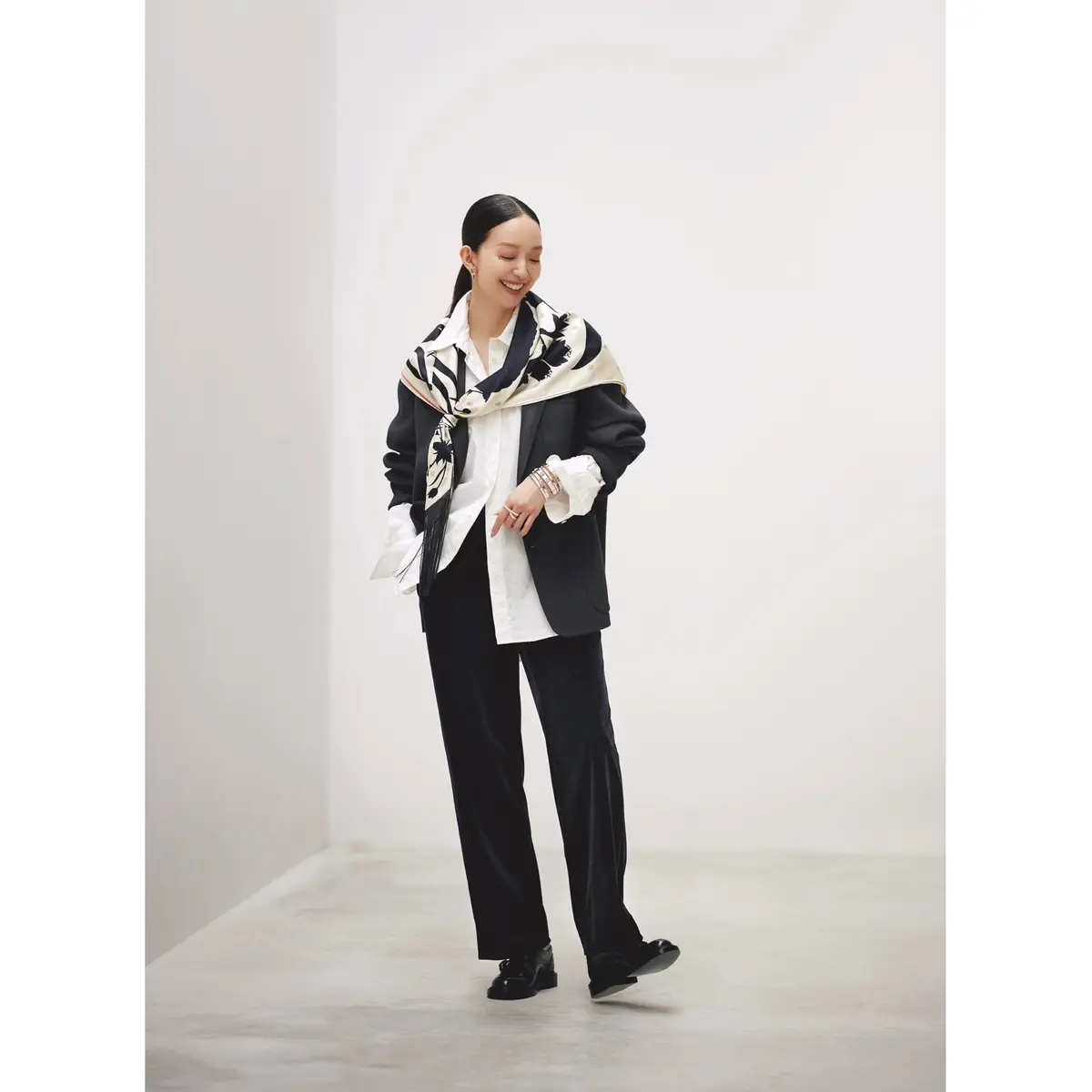 白シャツに黒ジャケットをはおり、肩から白×黒のプリントスカーフを巻き、黒のストレートパンツを合わせたコーディネート　モデル・松島花