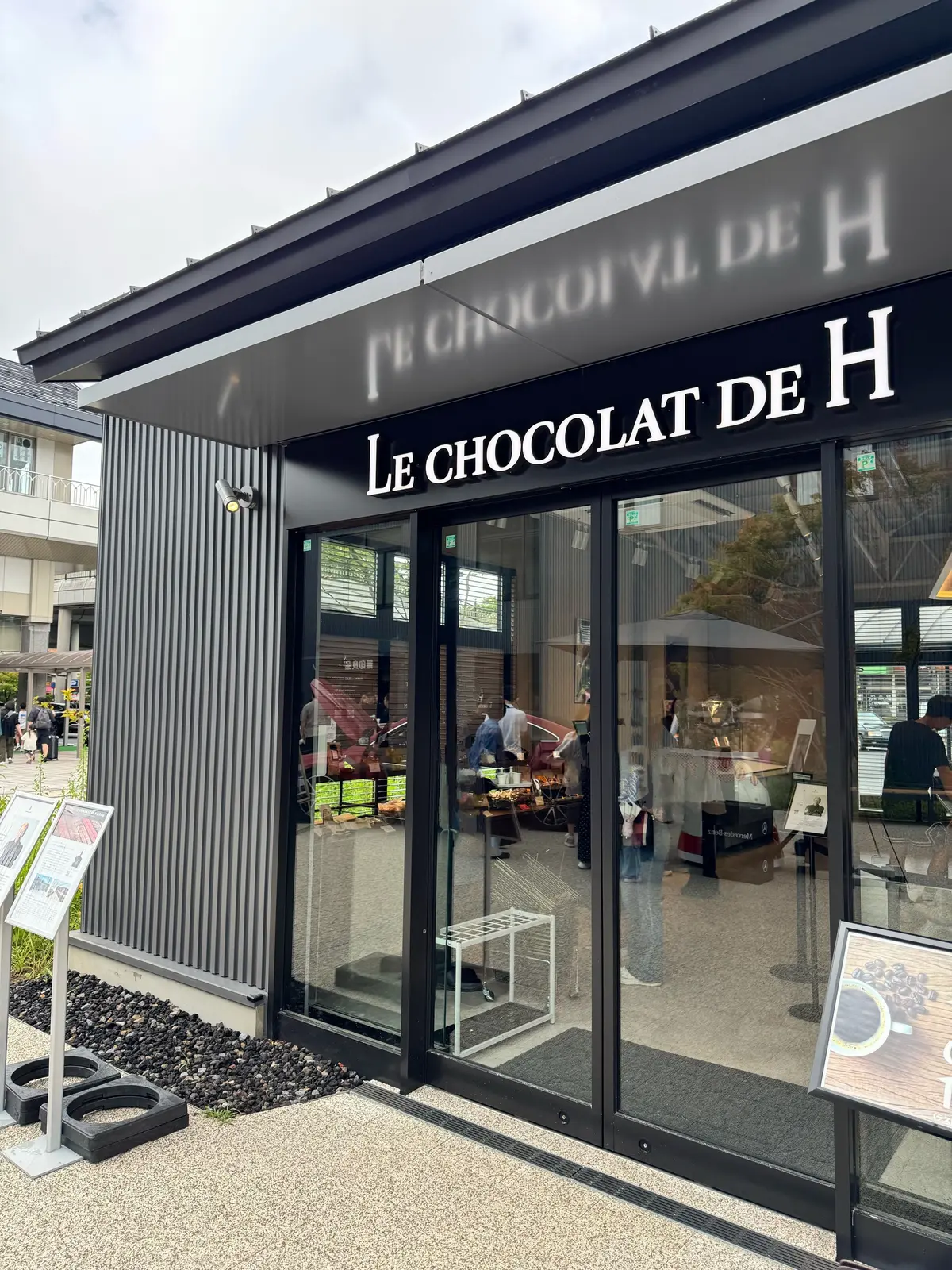 「ル・ショコラ・ド・アッシュ」のチョコレート店。グレーの建物で、大きなガラス窓に店内の商品が映り込んでいる。入り口前に白いベンチと石の敷き詰められた通路がある。