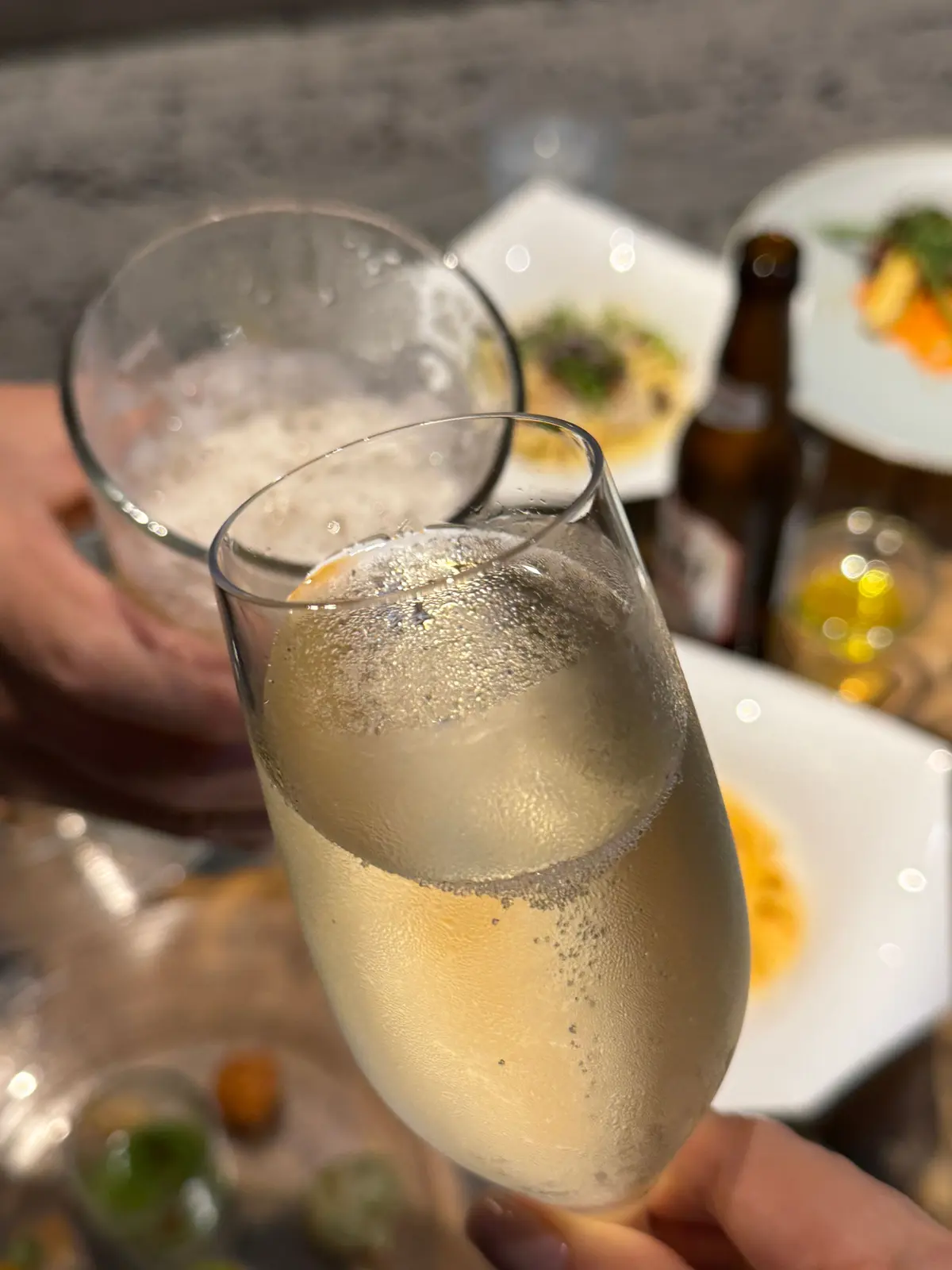 シャンパングラスに注がれた泡の立つ白ワイン。背景には料理とビール瓶が見える。テーブルで食事を楽しむ様子。