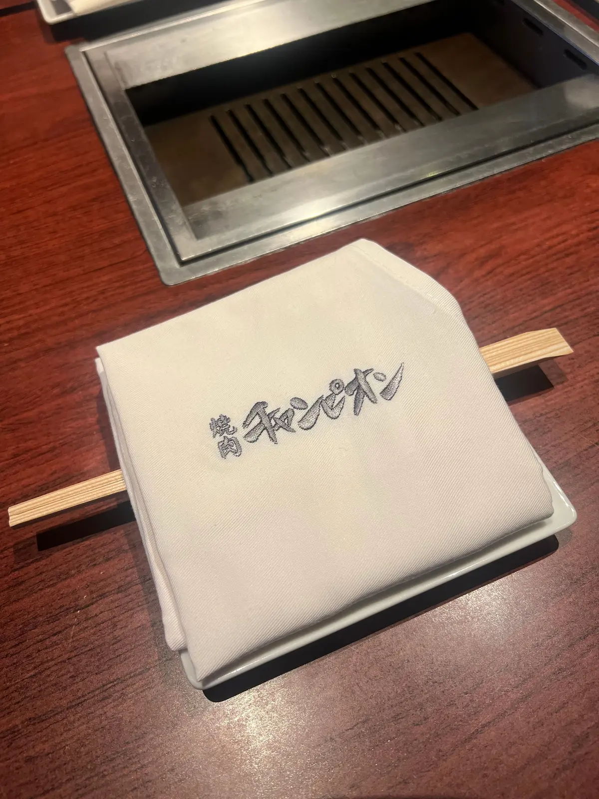 木製のテーブルに置かれた白いメニュー。メニューにはグレーの文字で「焼肉とジン」と書かれている。テーブルの上には、グリルが設置されている。