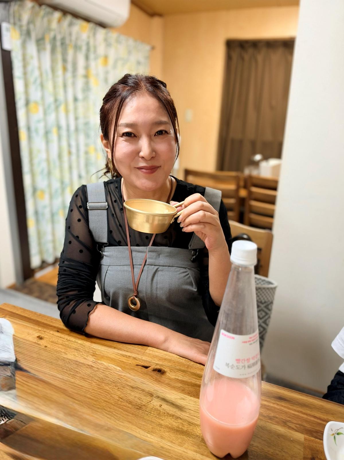 笑顔の女性が、木製のテーブルを挟んで、金色のカップとピンク色のボトルを手にしている。背景には、木製のドアと椅子が見える。