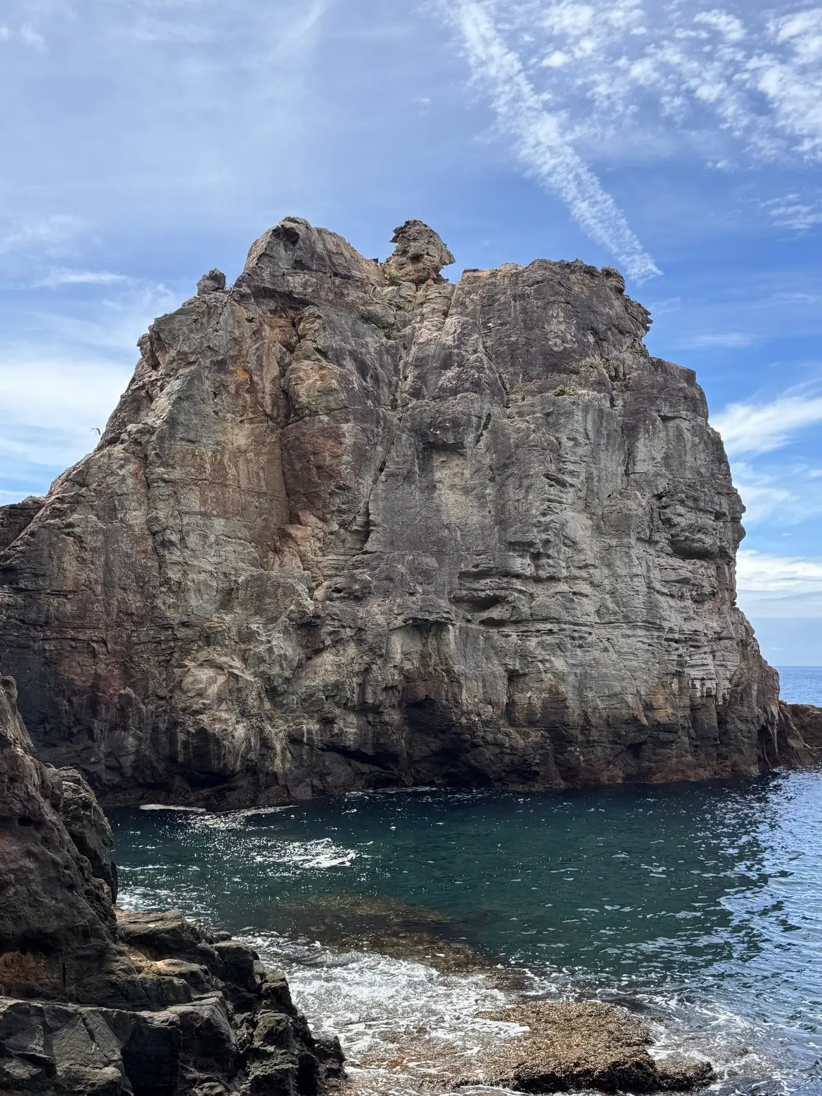 青い海に面した、灰色と茶色の層状の岩山。頂上付近に奇妙な形の岩がそびえ立つ。波が岩底に打ち寄せ、水面がキラキラと輝いている。