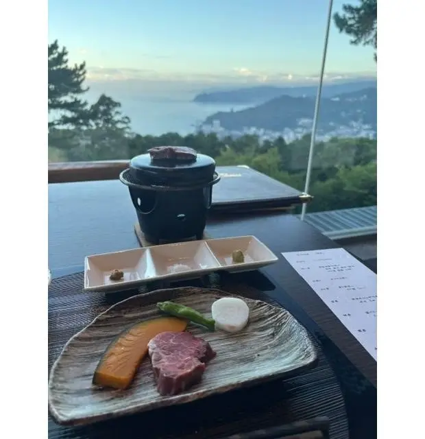 高台のレストランで、海と山を望む夕暮れ時の景色。テーブルには、陶器製の鍋料理、小皿に盛り付けられたお肉や野菜、薬味、調味料などが置かれている。