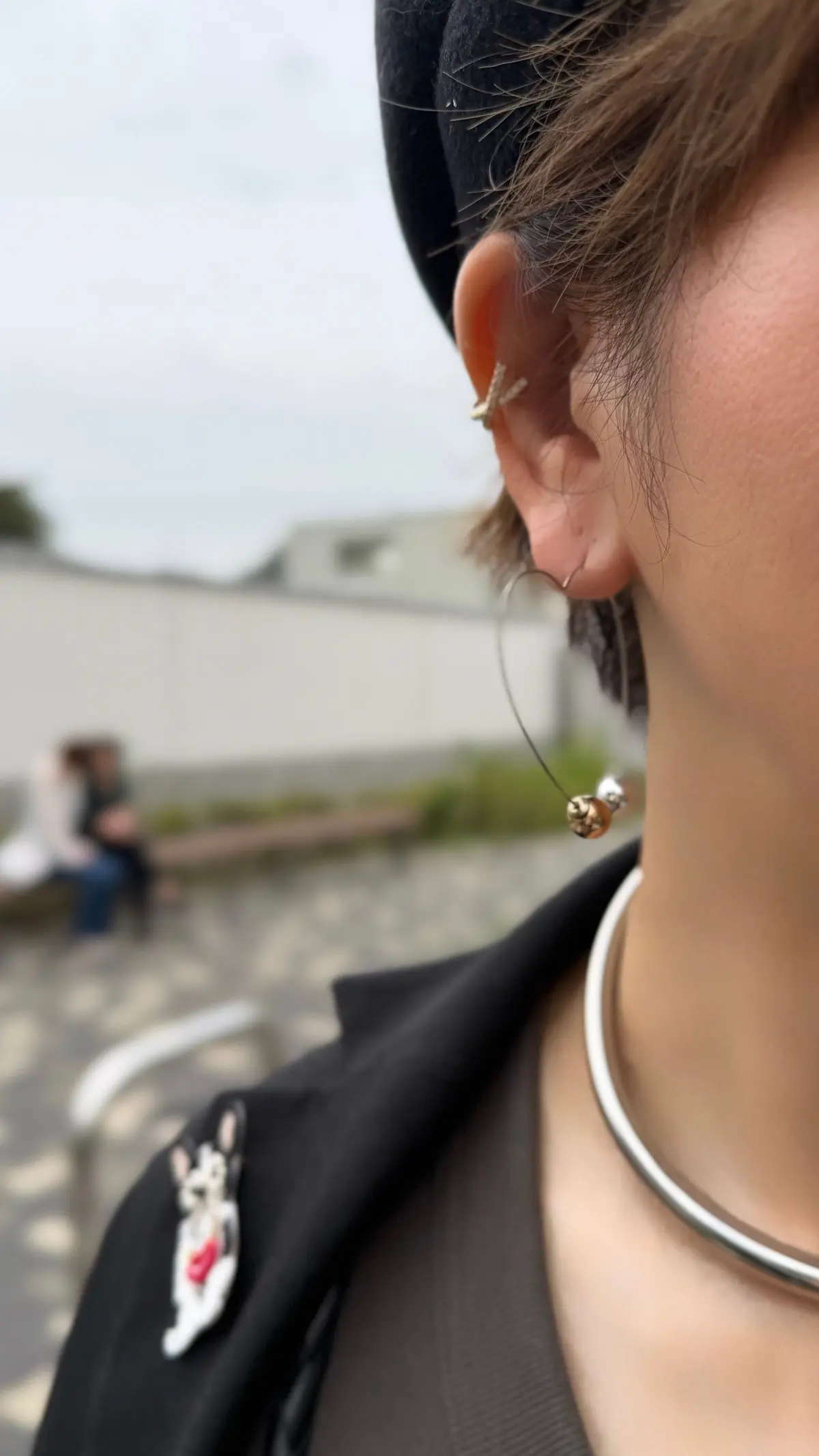 黒いニット帽を被った人物の耳元。片方の耳には金色の装飾が施されたスタッドピアス、もう片方の耳には銀色のリングピアスが垂らされている。首には銀色の太いチョーカーを着用。背景には現代的な建築物と人物がぼやけて見える。