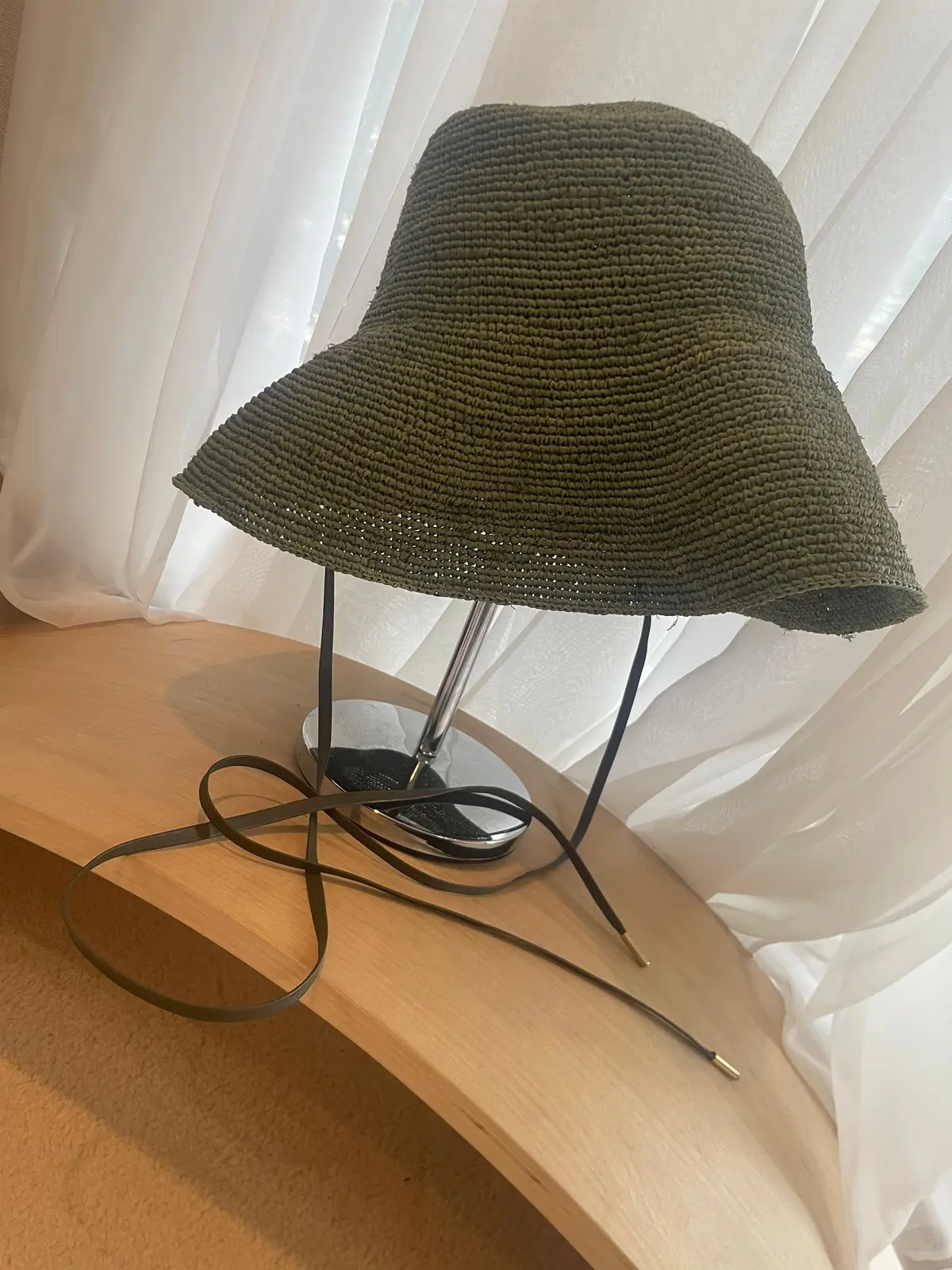 緑の編み帽子が木製のテーブルに置かれている。背景には白いカーテンが光を受けている。