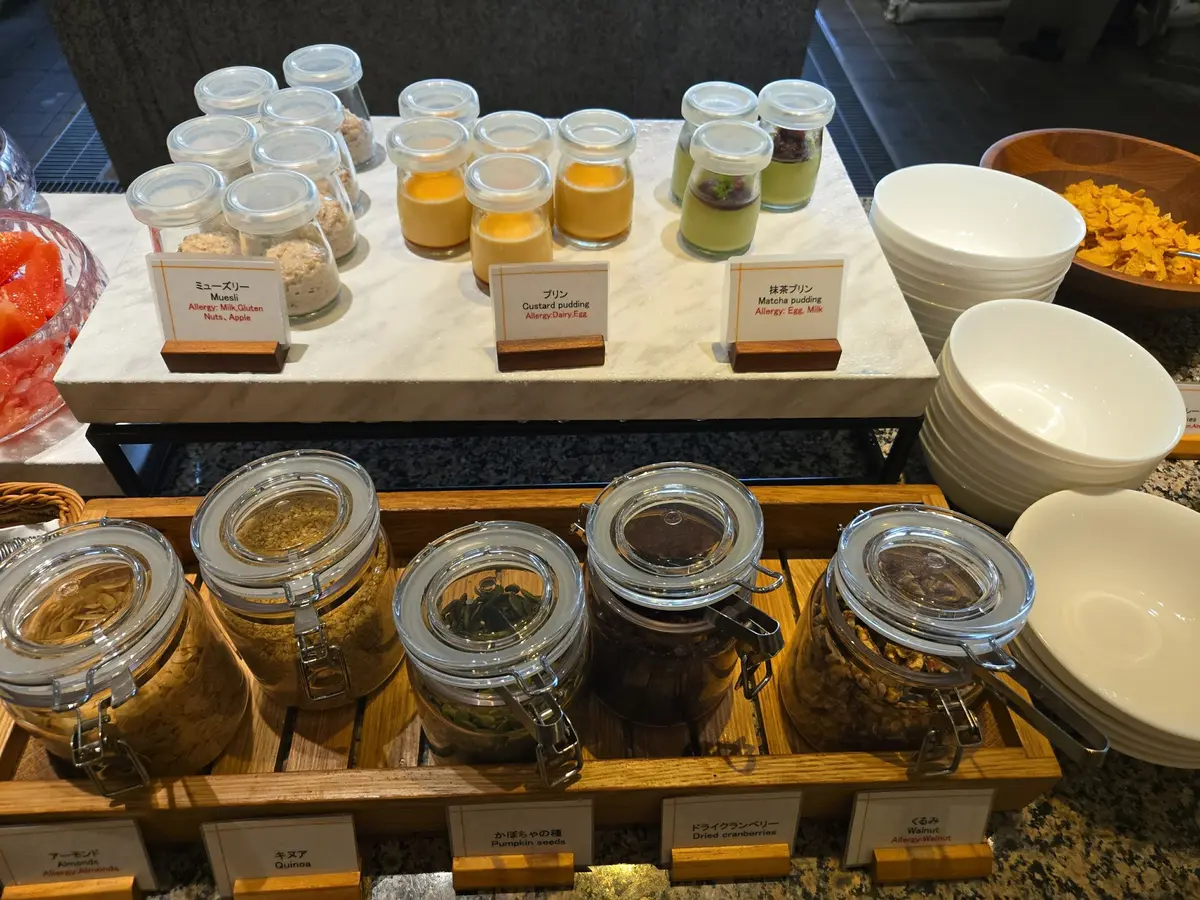 白い食器にフルーツやプリン、抹茶プリンが並び、下段には様々なナッツや種子がガラス容器に詰められているビュッフェの様子。日本語の食品アレルギー表示が添えられている。
