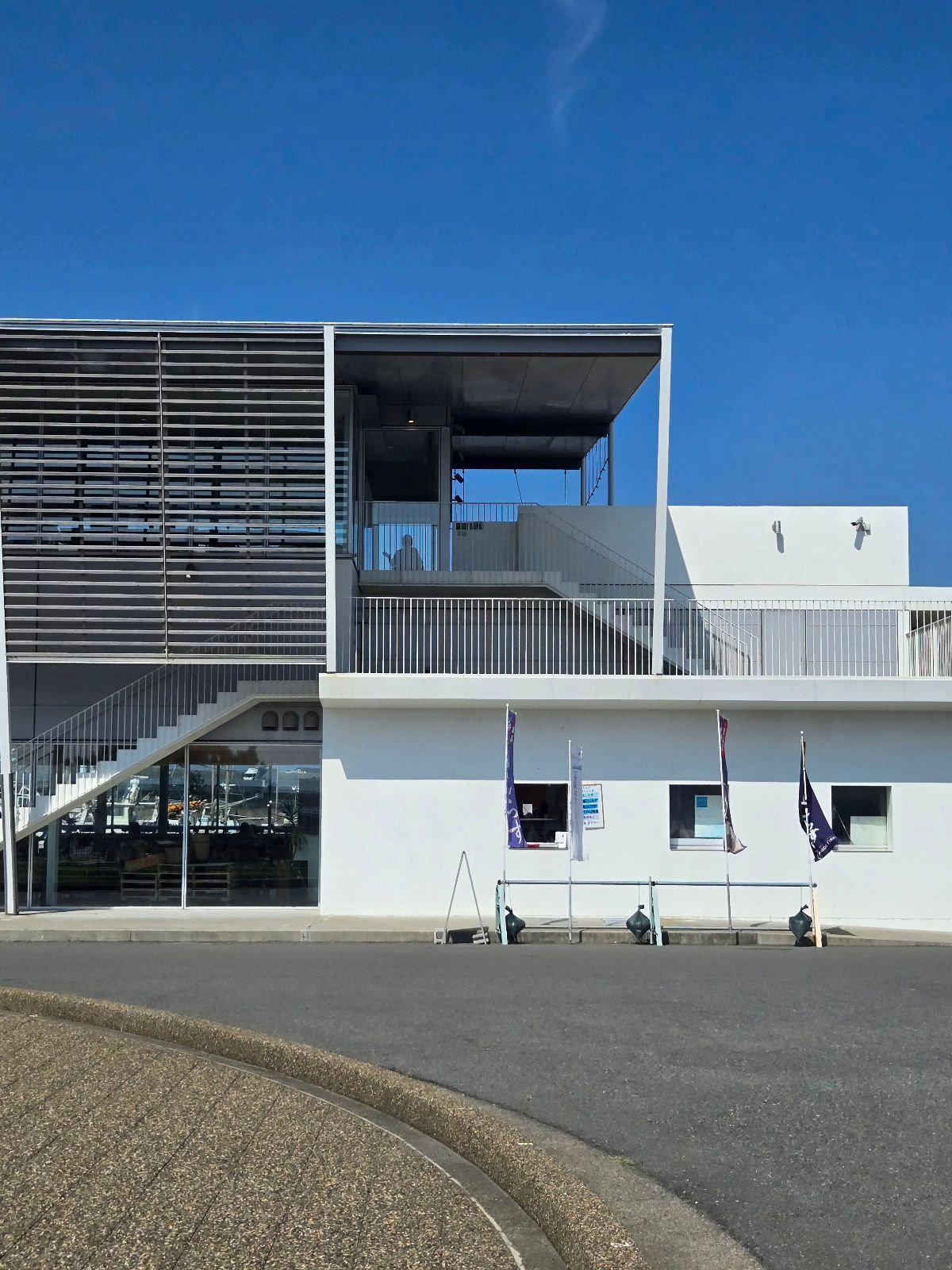 白い現代的な建物。正面に階段があり、上層階には屋根付きのテラスがある。旗が数本立てられている。背景は青空。