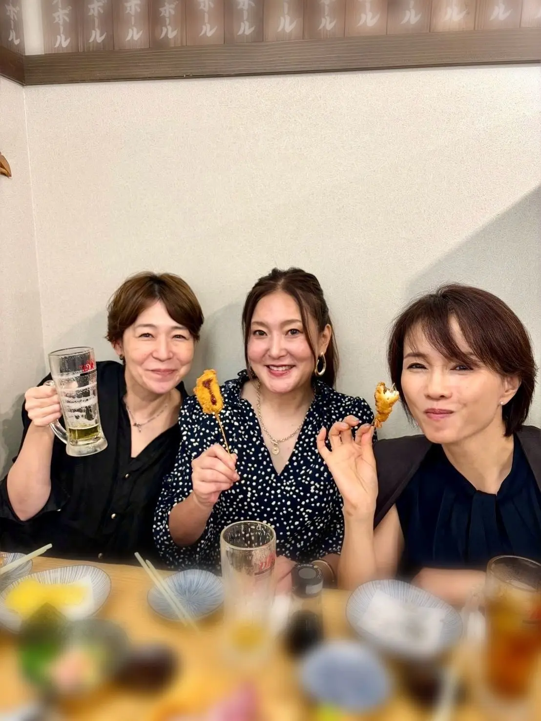 笑顔の女性3人が、日本料理店で食事を楽しんでいる様子。手にはビールや唐揚げを持ち、テーブルには料理や食器が並んでいる。背景には木製の装飾がある。